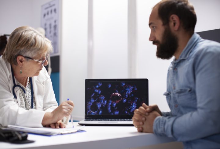 Old female physician and young male patient use laptop to analyze virus cells during consultation. Senior doctor with digital device illustrating enlarged microscopic details of germs to bearded man.